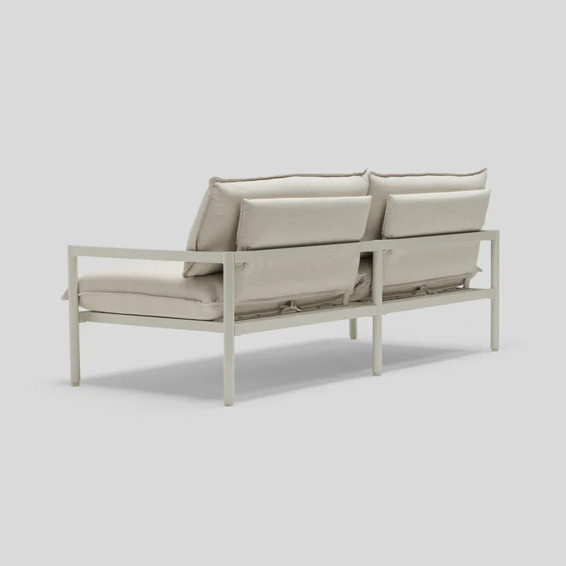 The Terra Loveseat is a minimalist outdoor sofa with a light metal frame and plush beige cushions, shown from the back angle. It features clean lines and a modern aesthetic, making it ideal for patio or garden settings.
