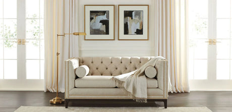 The Anderson Loveseat in beige with a throw blanket is placed against a backdrop of floor-to-ceiling curtains. Two abstract art pieces adorn the wall above the loveseat. To the left stands a tall, gold floor lamp, and dark wood flooring completes the room’s aesthetic.