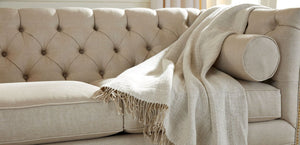 An Anderson Loveseat in beige, featuring tufted upholstery with a matching cushion, and a light fringed throw draped over one armrest, is set in a cozy living room environment.