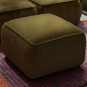A square, olive green corduroy ottoman from the Macy Modular Sofa collection sits on a textured red and blue striped rug. The ribbed texture of the ottoman's fabric gives it a plush and comfortable look, perfectly complementing the cozy aesthetic of the room.