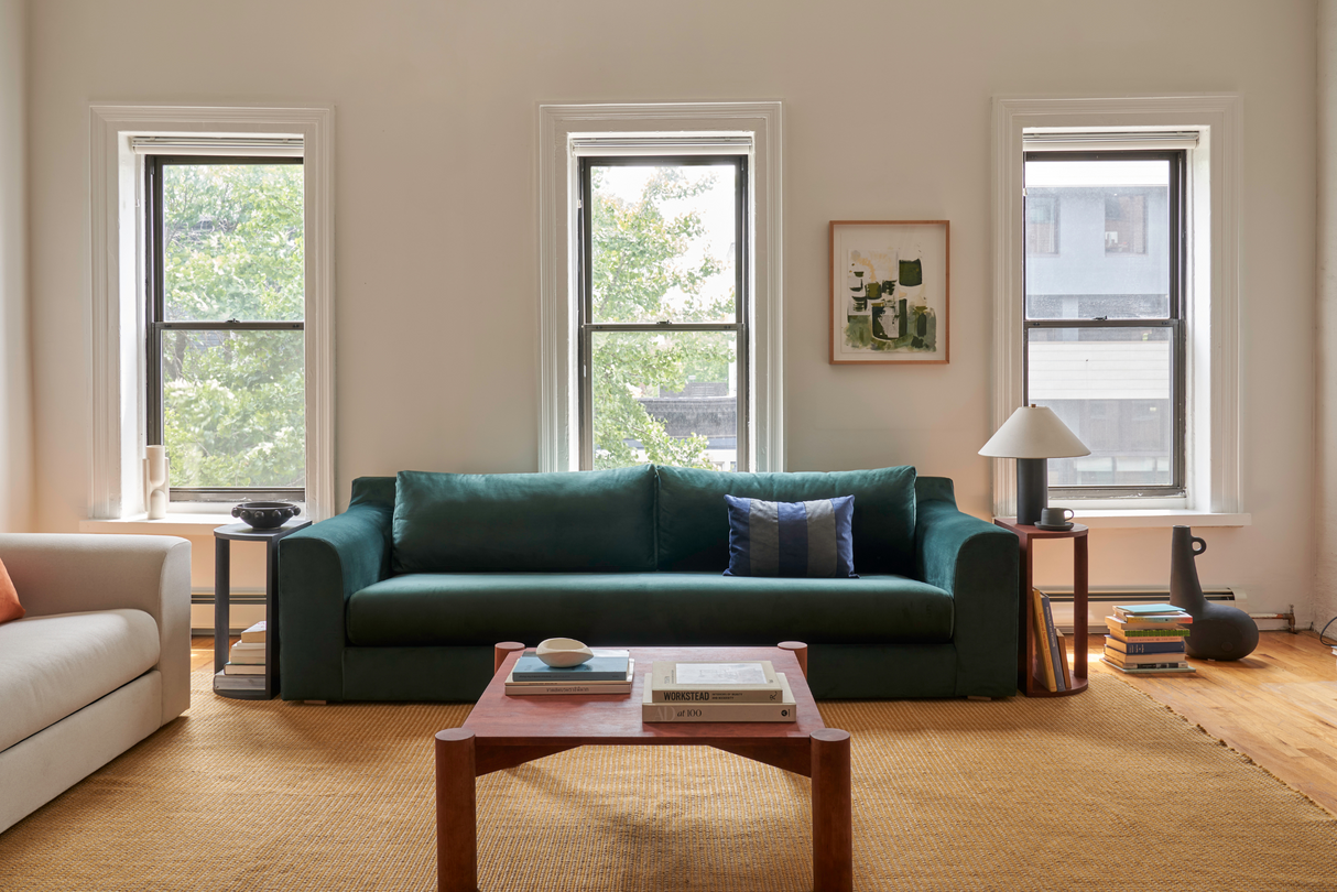 In a cozy living room, The Elevate Sofa in dark green is placed against a wall featuring three large windows. A wooden coffee table displays books and a decorative item. Beside the sofa, a modern lamp rests on a stack of books. A framed art piece is elegantly hung on the wall above.