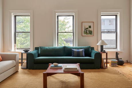 In a cozy living room, The Elevate Sofa in dark green is placed against a wall featuring three large windows. A wooden coffee table displays books and a decorative item. Beside the sofa, a modern lamp rests on a stack of books. A framed art piece is elegantly hung on the wall above.