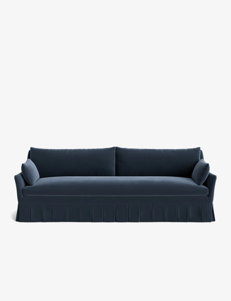 The Portola Pleated Sofa is a sleek, dark blue piece with a pleated skirt, featuring two back cushions and two bolster pillows on each end, all set against a plain white background.