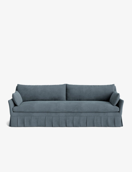 The Portola Pleated Sofa, in a teal velvet finish, showcases pleated skirt detailing, two large back cushions, and two cylindrical side pillows, all set against a plain white background.