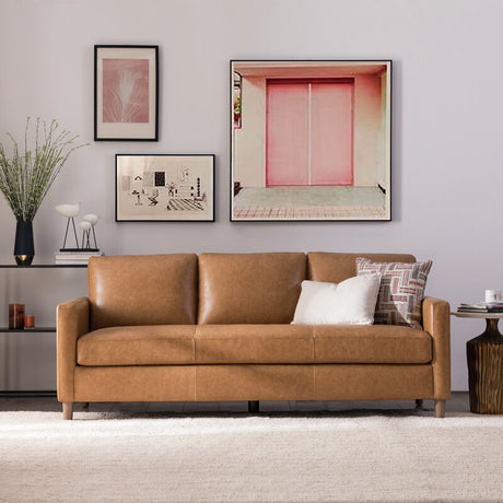 The living room exudes a cozy atmosphere with its Portland 80.5" Leather Sofa in brown, accented by cushions and accompanied by a side table and a potted plant. Above the sofa, four framed artworks are displayed on the wall, enhancing the decorative appeal of the neutral-toned space.