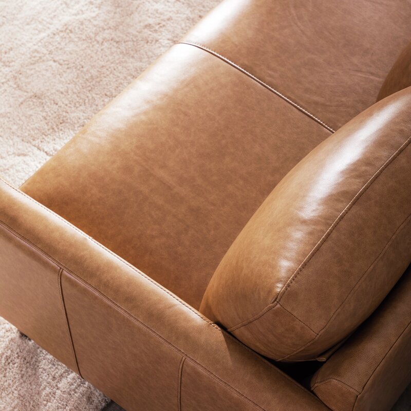A close-up of the Portland 80.5" Leather Sofa highlights its brown leather upholstery and plush cushions, set against a light beige carpet. The smooth texture and detailed stitching of the leather emphasize the sofa's quality craftsmanship.