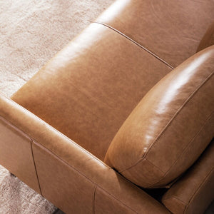 A close-up of the Portland 80.5" Leather Sofa highlights its brown leather upholstery and plush cushions, set against a light beige carpet. The smooth texture and detailed stitching of the leather emphasize the sofa's quality craftsmanship.