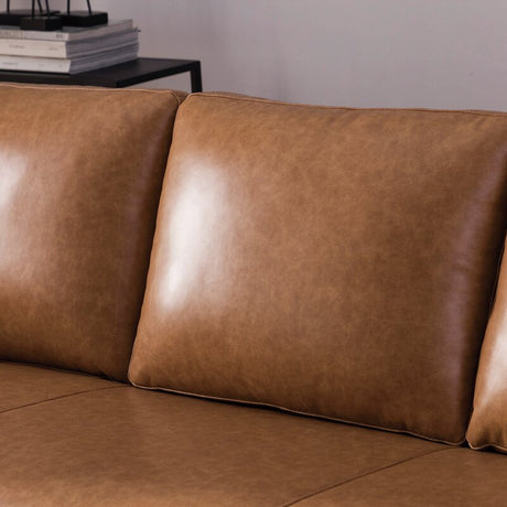 Close-up of the Portland 80.5" Leather Sofa in rich brown leather with plush cushions. The image highlights the sofa's smooth texture and sleek design, complemented by a stack of books in the background on a black side table.