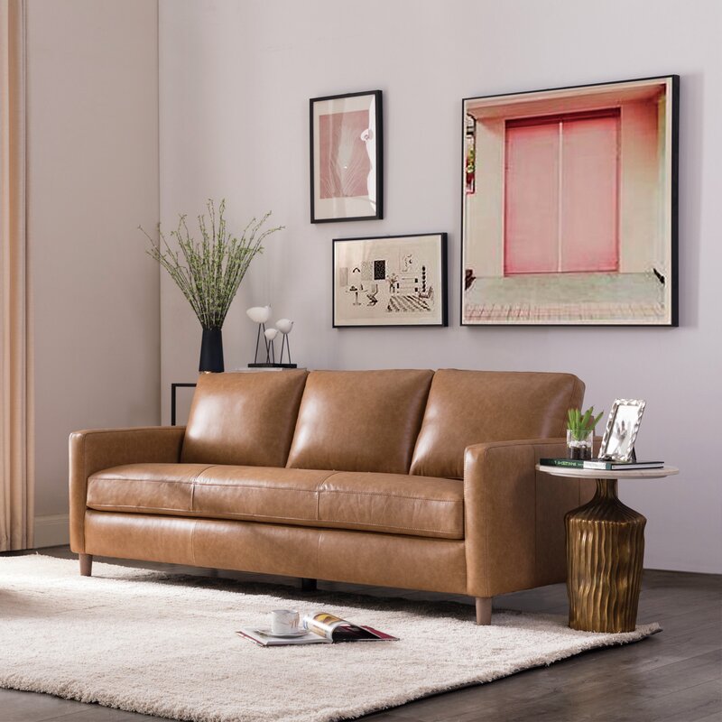 A modern living room featuring the Portland 80.5" Leather Sofa in brown, resting on a light rug. A small side table holding books, a plant, and two glasses is positioned beside the sofa. Three artworks in black frames adorn the white wall, while natural light streams in from a window on the left.