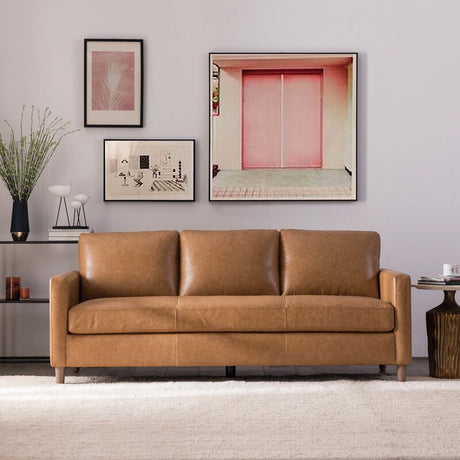 A cozy living room features the Portland 80.5" Leather Sofa in tan, placed on a light rug. Behind the sofa, a wall is adorned with framed art showcasing abstract designs and a pink-toned photograph. Beside the sofa is a small table with a vase filled with green branches.