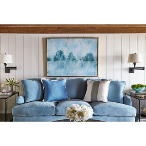 A cozy living room features the Brooke 6-Seat Sofa adorned with pillows in shades of blue and beige. An abstract blue painting hangs on a white shiplap wall above the sofa. Side tables showcase plants and lamps, while a vase with flowers rests on the coffee table.