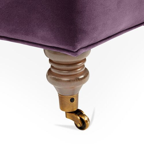 Close-up of a Brooke 6-Seat Sofa leg with a decorative wooden spindle design, featuring a small brass caster at the bottom. The fabric above the leg is a deep purple color.