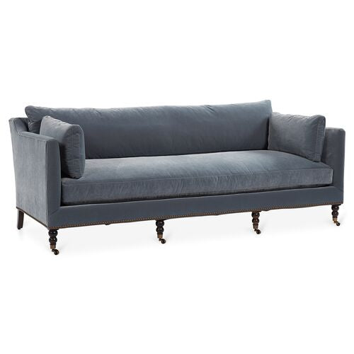 The Margot Velvet Sofa is a gray three-seater with rolled armrests, nailhead trim, and turned wooden legs with casters. Its design is elegant and classic, making it suitable for a living room setting.