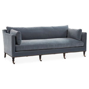The Margot Velvet Sofa is a gray three-seater with rolled armrests, nailhead trim, and turned wooden legs with casters. Its design is elegant and classic, making it suitable for a living room setting.