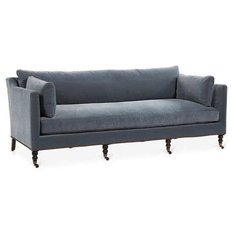 The Margot Velvet Sofa is a gray three-seater with rolled armrests, nailhead trim, and turned wooden legs with casters. Its design is elegant and classic, making it suitable for a living room setting.