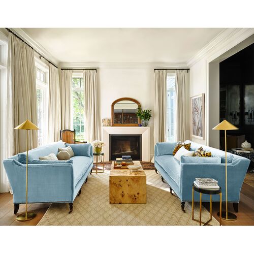 A cozy living room featuring two blue Margot Velvet Sofas facing each other, with a wooden coffee table in between, and a fireplace adorned with a large mirror above it. Tall windows with light curtains and gold floor lamps flank either side, while a patterned rug graces the wooden floor.