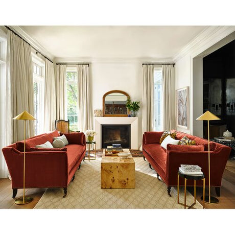 The inviting living room features two Margot Velvet Sofas in a rich red color facing each other, with a wooden coffee table placed between them. A fireplace is centrally positioned between the sofas, topped with a mirror above it. Tall lamps and large windows adorned with sheer curtains beautifully accentuate the space.
