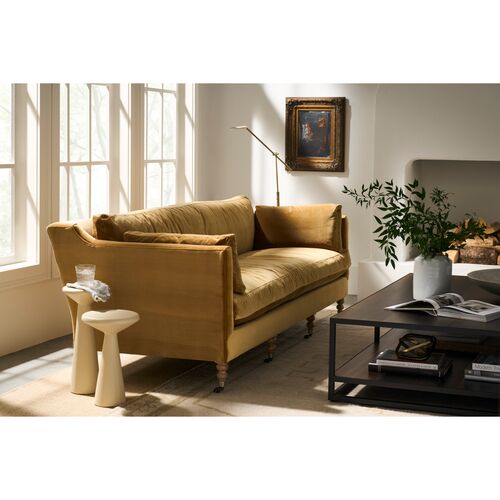 A cozy living room features the Margot Velvet Sofa in mustard-yellow by a large window. A small side table holds a glass of water, while a painting is displayed above the sofa. In front of it, a coffee table with an open book and a potted plant completes the setting.