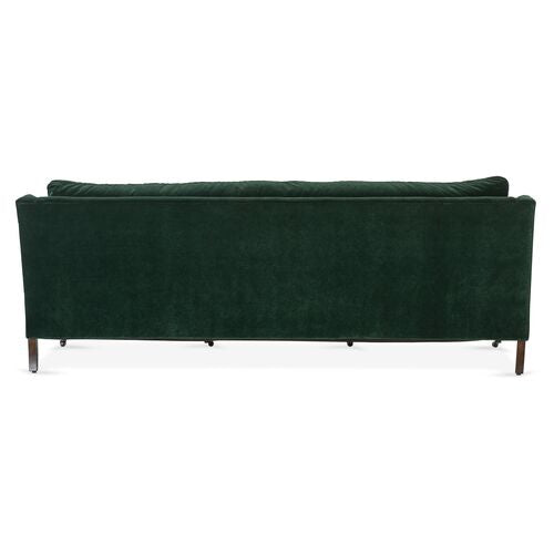 A Margot Velvet Sofa in dark green velvet is showcased from the back, highlighting its sleek, rectangular design and wooden legs. The image captures the sofa in a well-lit environment against a simple background.