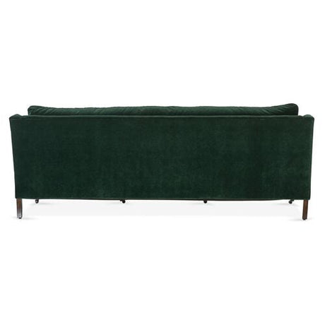 A Margot Velvet Sofa in dark green velvet is showcased from the back, highlighting its sleek, rectangular design and wooden legs. The image captures the sofa in a well-lit environment against a simple background.