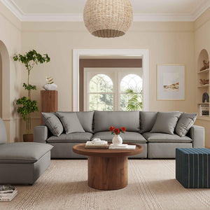 A cozy living room features the Cali Performance Fabric Modular 4 Piece Sectional in gray, paired with a round wooden coffee table. A large wicker pendant light hangs overhead, while decor includes a potted plant, an assortment of books, and a vase filled with red flowers. Neutral tones and natural light contribute to the warm atmosphere.
