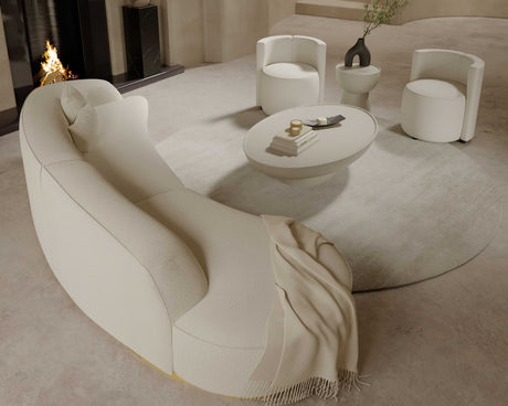 A modern living room featuring the Reya Curved Sectional in beige, accompanied by two white armchairs and a round coffee table placed on a circular rug. A fireplace serves as a backdrop, while a beige throw is elegantly draped over the sectional. A vase with branches adorns the table.