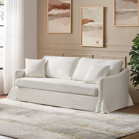 The Torsten Modern Comfortable Sofa, which is white and comes with three cushions, is positioned against a beige wall adorned with two abstract paintings. On the left, there's a large window draped with sheer curtains, while a green plant accents the right side. The floor showcases a light-colored patterned rug.