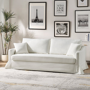 A modern living room showcases the Torsten Modern Comfortable Sofa in white, topped with two cushions, and paired with a light-colored rug. The wall behind displays framed art, featuring a picture of the Eiffel Tower. To the left stands a potted plant, while on the right, a sideboard is partially visible.