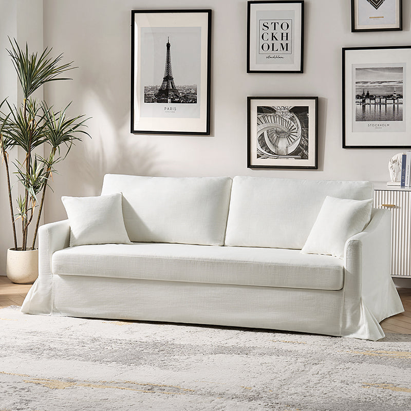 A modern living room showcases the Torsten Modern Comfortable Sofa in white, topped with two cushions, and paired with a light-colored rug. The wall behind displays framed art, featuring a picture of the Eiffel Tower. To the left stands a potted plant, while on the right, a sideboard is partially visible.