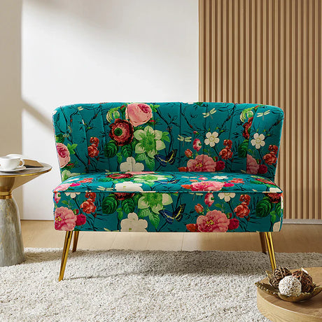 The Coraline Loveseat, featuring a vibrant floral pattern with gold legs set against a teal background, is placed on a white rug. Next to it stands a small round table adorned with a decorative item. The room is accentuated by wooden panel walls and flooring, illuminated by streaming sunlight.