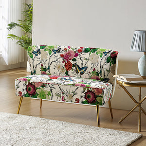 The Coraline Loveseat, featuring a vibrant floral and butterfly design, rests elegantly on a light wooden floor. Beside it stands a small round table adorned with a blue lamp and a book. Sunlight pours through sheer curtains, casting light onto the scene as a potted plant adds greenery to the background.