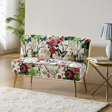 The Coraline Loveseat, featuring a vibrant floral and butterfly design, rests elegantly on a light wooden floor. Beside it stands a small round table adorned with a blue lamp and a book. Sunlight pours through sheer curtains, casting light onto the scene as a potted plant adds greenery to the background.
