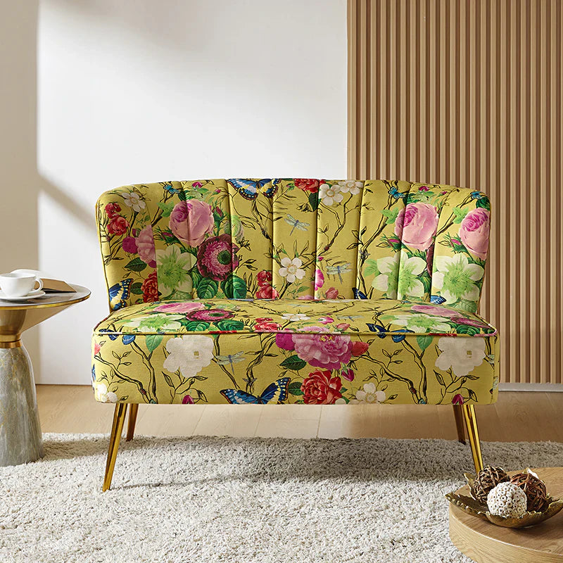 The Coraline Loveseat is a small, elegant piece adorned with a yellow floral and butterfly pattern, showcasing roses and green foliage. It features gold legs and is situated in a modern room with wooden flooring and a wooden panel accent wall. Adjacent to it is a round side table holding a cup and saucer.