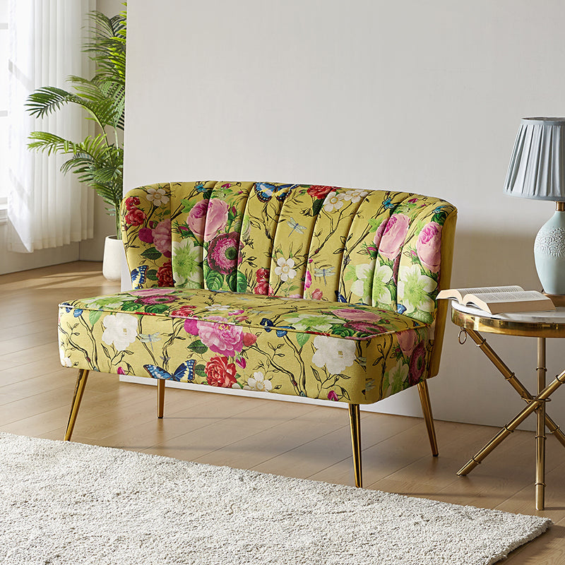 The Coraline Loveseat, showcasing a vivid floral design adorned with roses and butterflies, sits on a light wood floor. Beside it stands a round table with a lamp, located near a large window draped with sheer white curtains and accompanied by a potted plant. A white rug complements the floor setting.