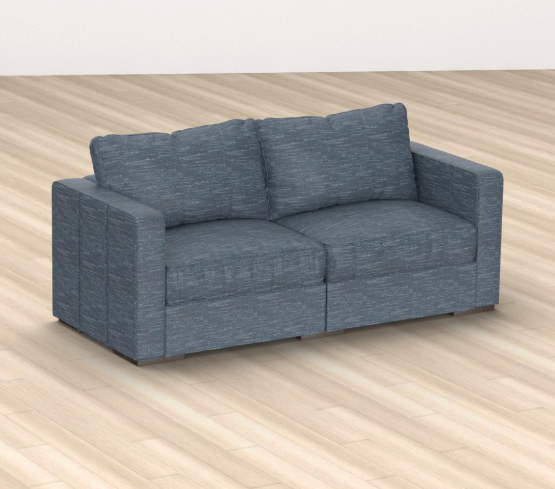 A vintage blue loveseat with a rained chenille fabric sits on a light wooden floor. The Sactionals sofa features square armrests and two seat cushions, complemented by two back cushions. The setting is minimalistic and modern.