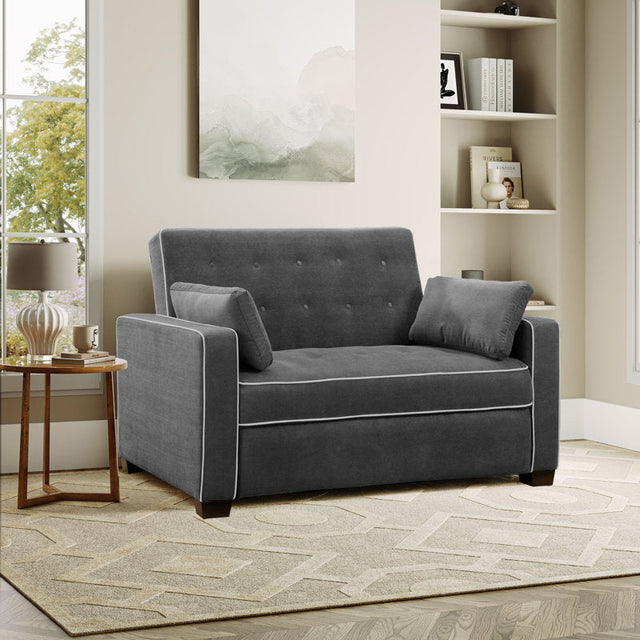 A cozy living room showcasing the Serta Monroe Full Size Convertible Sofa in a dark gray finish with white piping, complemented by two matching cushions. It rests on a beige patterned rug. A window offers a view of trees, and a bookshelf displays decorative items and books.