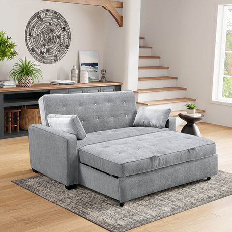 A modern living room showcases the Serta Monroe Full Size Convertible Sofa, featuring a gray, tufted design with plush cushions. The sofa rests on a patterned rug and is complemented by a small black side table nearby. A wooden staircase leads upward, while a decorative wall piece enhances the light-colored wall.