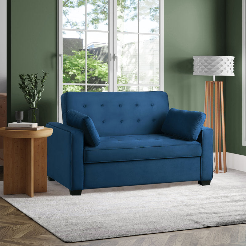 A cozy living room features the Serta Monroe Full Size Convertible Sofa in blue, complete with a tufted backrest and two bolster cushions. It rests on a light gray rug, positioned next to a round wooden side table adorned with a plant. A large window offers views of greenery, while a standing lamp is conveniently situated nearby.