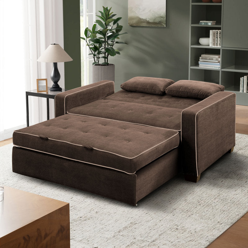 A Serta Monroe Full Size Convertible Sofa in brown is unfolded in a bright living room. It features two cushions, complemented by a small wooden side table with a lamp, a potted plant, and shelves in the background. The decor boasts modern elements with neutral tones.