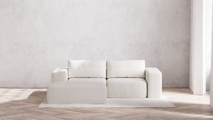 A minimalist living room showcasing a modern Modular 2-Seater Chaise Sectional in Boucle Aspen, complete with square cushions and a matching ottoman. The arrangement sits on a light-colored, textured rug over wooden flooring, with gentle natural light streaming from a window to the right.