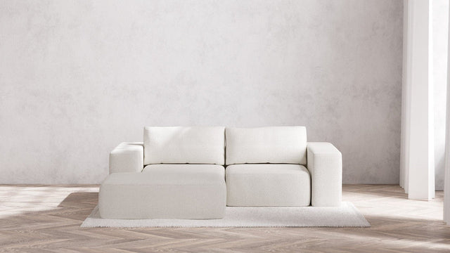 A minimalist living room showcasing a modern Modular 2-Seater Chaise Sectional in Boucle Aspen, complete with square cushions and a matching ottoman. The arrangement sits on a light-colored, textured rug over wooden flooring, with gentle natural light streaming from a window to the right.