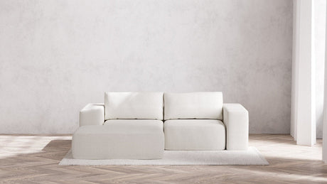 A minimalist living room showcasing a modern Modular 2-Seater Chaise Sectional in Boucle Aspen, complete with square cushions and a matching ottoman. The arrangement sits on a light-colored, textured rug over wooden flooring, with gentle natural light streaming from a window to the right.