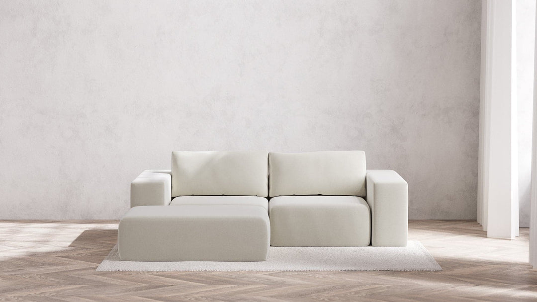 A minimalist living room showcases a Modular 2-Seater Chaise Sectional in Boucle Aspen with an accent ottoman on a textured rug. The backdrop consists of a plain, light gray wall, while sunlight softly illuminates the wooden herringbone floor with gentle shadows.