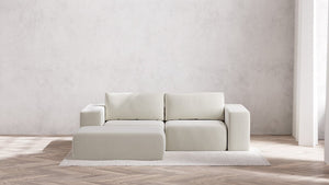 A minimalist living room showcases a Modular 2-Seater Chaise Sectional in Boucle Aspen with an accent ottoman on a textured rug. The backdrop consists of a plain, light gray wall, while sunlight softly illuminates the wooden herringbone floor with gentle shadows.