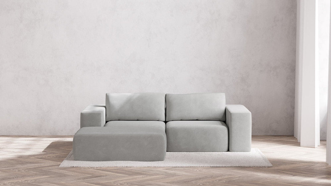 A minimalistic living room with a Modular 2-Seater Chaise Sectional in Boucle Aspen, featuring plush cushions and a matching ottoman. The space is bright, filled with soft natural light and surrounded by neutral-colored walls, complemented by a light herringbone wooden floor and a simple rug.