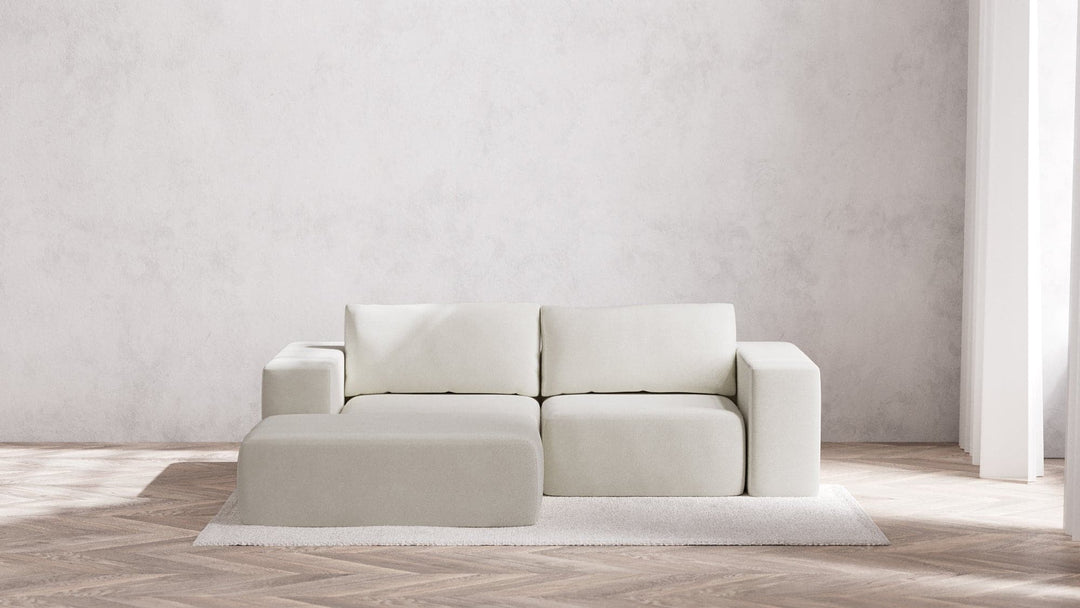 A minimalist living room showcases the Modular 2-Seater Chaise Sectional in Boucle Aspen, accompanied by an ottoman on a textured area rug. This setting includes a herringbone wood floor and a plain white wall that fosters a clean and serene atmosphere, with natural light streaming through the tall window.