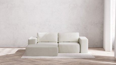 A minimalist living room showcases the Modular 2-Seater Chaise Sectional in Boucle Aspen, accompanied by an ottoman on a textured area rug. This setting includes a herringbone wood floor and a plain white wall that fosters a clean and serene atmosphere, with natural light streaming through the tall window.