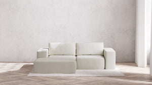 A minimalist living room showcases the Modular 2-Seater Chaise Sectional in Boucle Aspen, accompanied by an ottoman on a textured area rug. This setting includes a herringbone wood floor and a plain white wall that fosters a clean and serene atmosphere, with natural light streaming through the tall window.