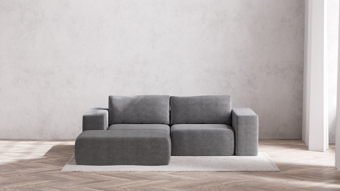 A chic Modular 2-Seater Chaise Sectional in Boucle Aspen sits atop a light-colored rug on a wooden herringbone floor. The minimalist decor includes a simple white wall bathed in generous natural light from unseen windows.