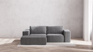 A chic Modular 2-Seater Chaise Sectional in Boucle Aspen sits atop a light-colored rug on a wooden herringbone floor. The minimalist decor includes a simple white wall bathed in generous natural light from unseen windows.